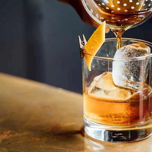 A whiskey on the rocks being poured, with a lemon twist garnish and a wine glass overhead. (140 chars, ends with a period)