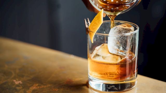 A whiskey on the rocks being poured, with a lemon twist garnish and a wine glass overhead. (140 chars, ends with a period)