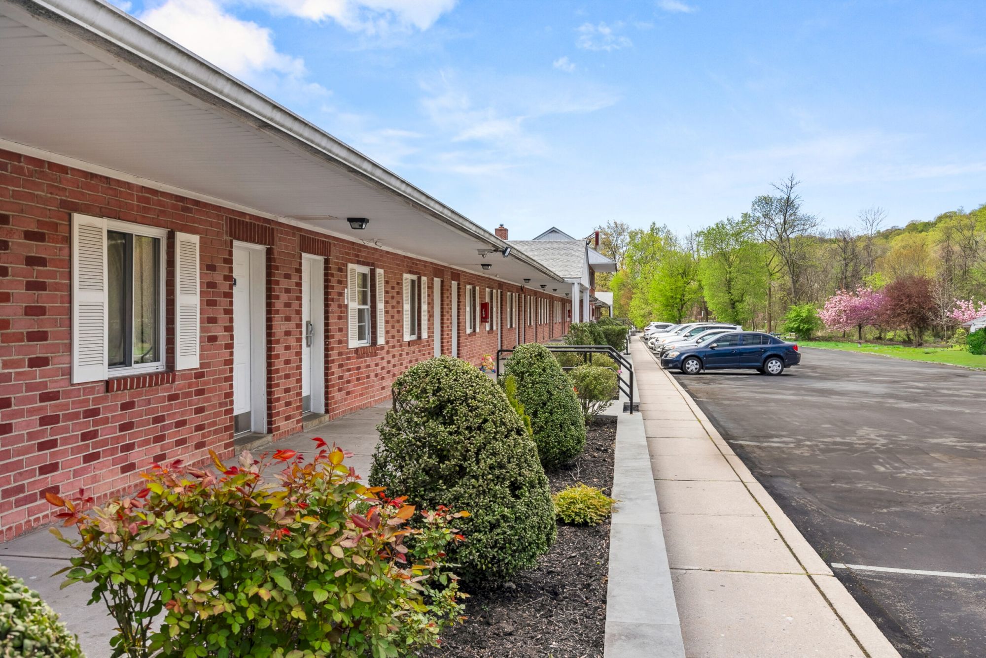 A single-story red brick motel-style building with white doors, a paved sidewalk, shrubs, and a parking lot with a blue car in a sunny, tree-lined lot.