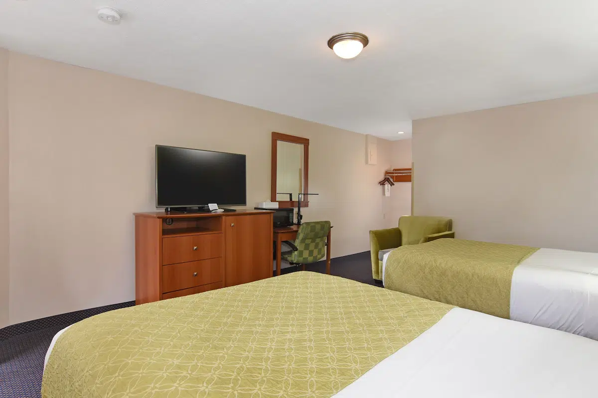 A tidy hotel room with two beds, a flat-screen TV on a dresser, and green chairs under ceiling lights.