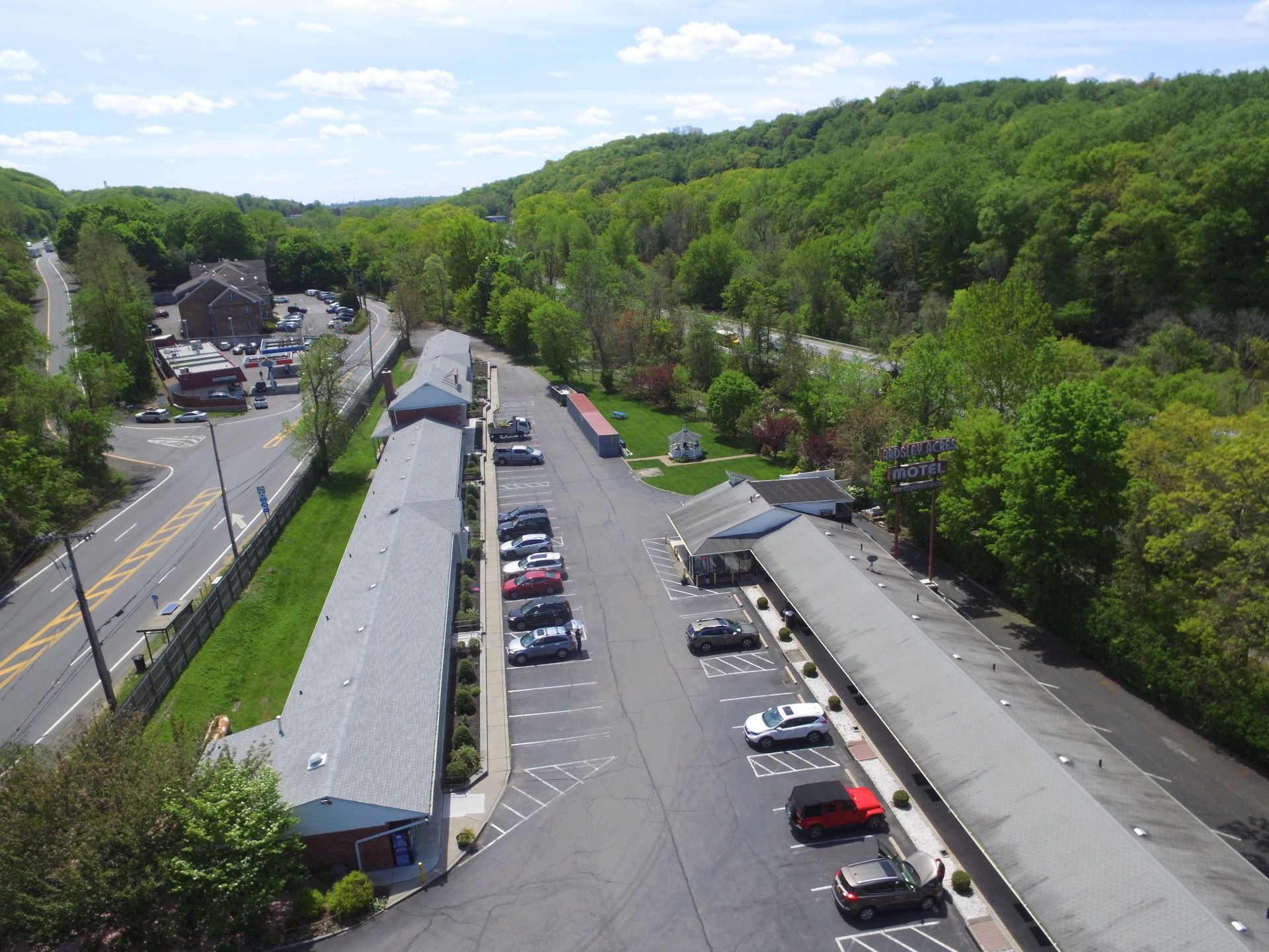 Aerial view of a small motel and row of cars parked in the lot, surrounded by green trees along a highway in a peaceful suburban area.