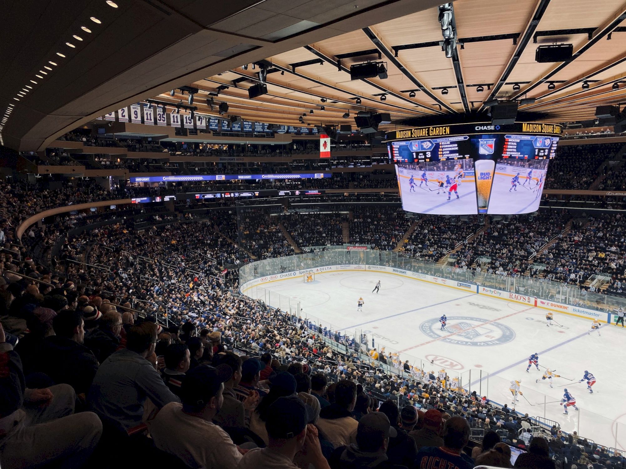 A busy hockey arena filled with spectators watching a game on the ice, with a large scoreboard and players in action.