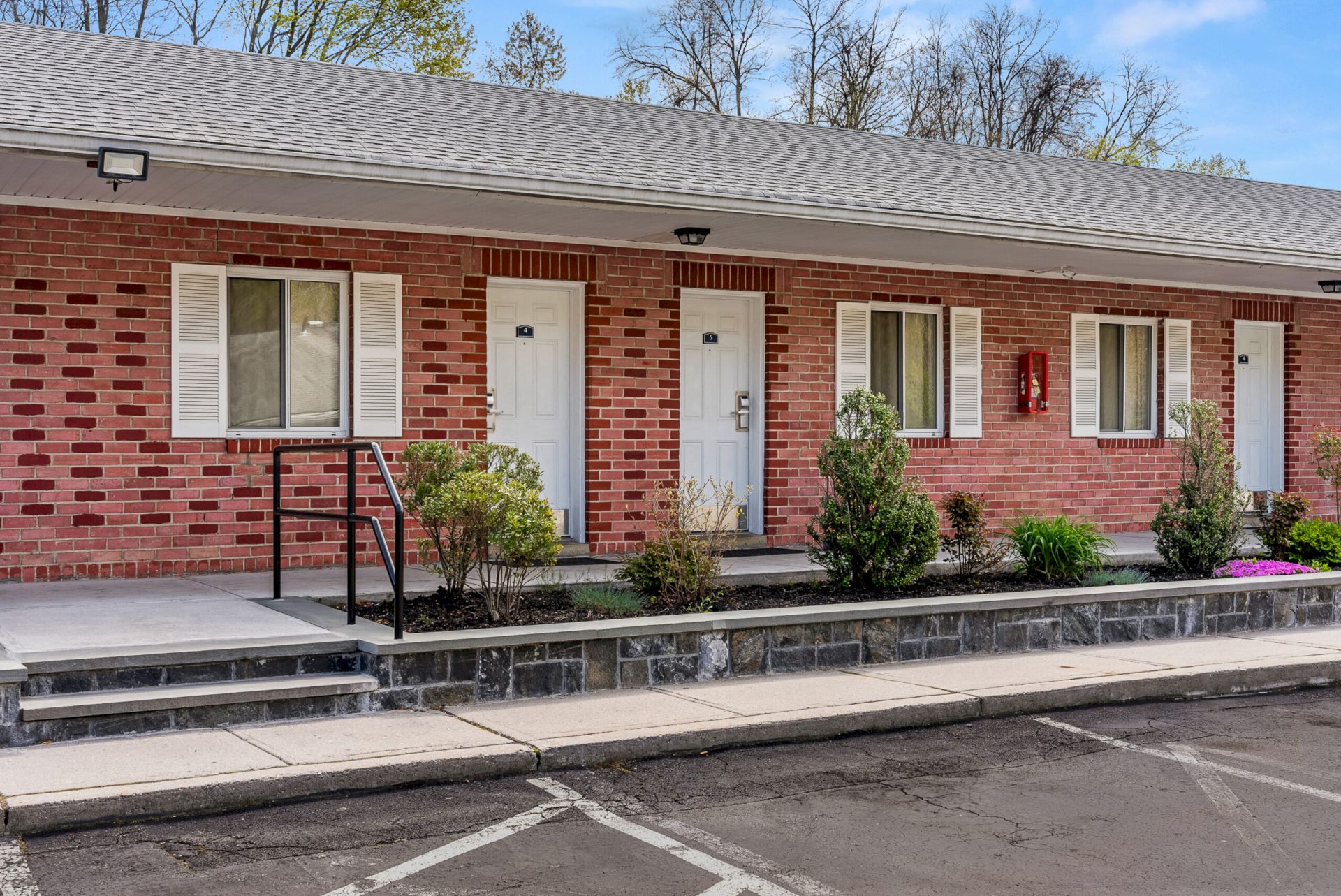 A red-brick building with white doors and shutters, windows, landscaped shrubs, and an outdoor fire extinguisher mounted on the wall.