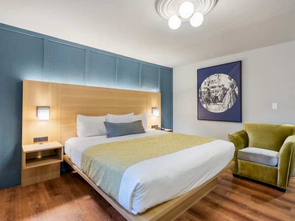 A modern hotel room with a large bed, blue wall paneling, wooden headboard, mustard throw, two bedside lamps, a lime-green armchair, and a framed artwork on the wall, all on wooden flooring.