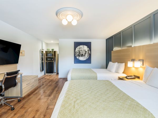 A bright hotel room with two double beds, a blue accent wall, a wall-mounted TV, a desk and chair, and modern lighting.