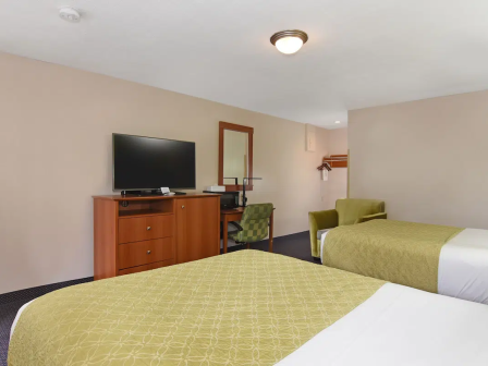 Two beds with green bed runners, a dresser with a flat-screen TV, a desk with a chair, and a couple of green armchairs in a hotel room.