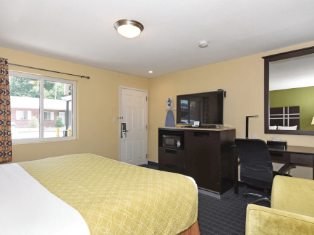 A hotel room with a queen bed, yellow bedspread, a window with patterned curtains, a desk area with a TV, and a dark wood dresser in a neat, yellow-walled space.