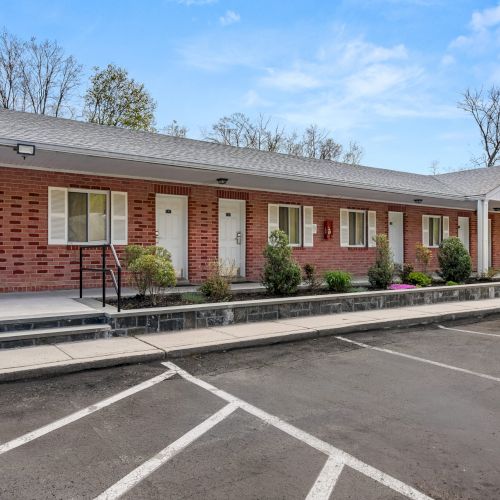 A row of single-story brick motel rooms with doors and windows, small shrubs along the walkway, a parking lot in front, and a clear blue sky.