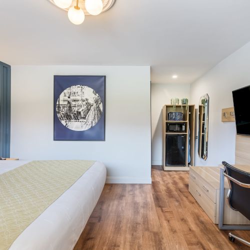 A clean hotel room with a bed, blue accent wall, wall art, wooden floor, desk with chair, and a large wall-mounted TV.