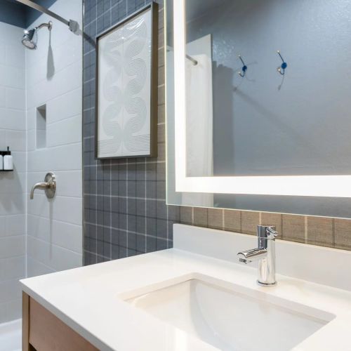 A modern bathroom with a white sink, sleek faucet, backlit mirror, gray tiled wall, and a shower area with a small shelf and bottles.