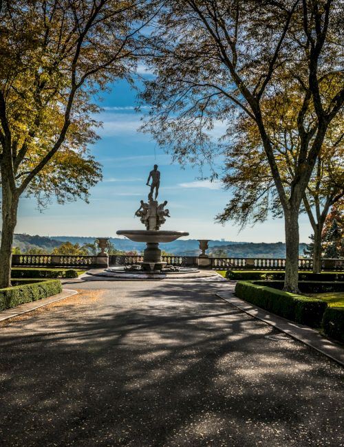 A formal garden with a central fountain, trimmed hedges, and tall trees framing a view toward the horizon by the water, under a sunny sky.